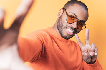 Wall Mural - Smiling dark-skinned young man wearing sunglasses taking a photo, selfie self-portrait with mobile phone while making victory gesture, isolated over orange background.