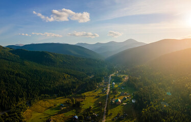 Wall Mural - Aerial view of amazing scenery with small village houses between foggy dark mountain peaks covered with forest pine trees at autumn sunrise. Beautiful wild woodland with shining rays of light at dawn