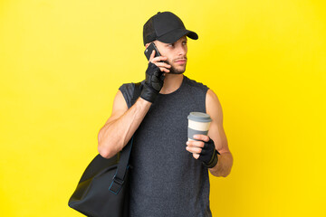 Wall Mural - Young sport blonde man with sport bag isolated on yellow background holding coffee to take away and a mobile