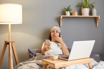 indoor shot of sleepy tired woman wearing sleep mask sitting with laptop in bed at home, holding mob