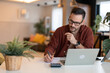 © Dorde - Serious millennial man using laptop sitting at the table in a home office. Focused guy in casual clothing looking at the paper, writing down ideas or a future business plan and using laptop.