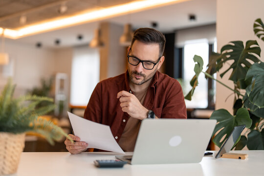 Serious millennial man using laptop sitting at the table in a home office, looking at the paper, communicating online, writing emails, distantly working or studying on computer at home.