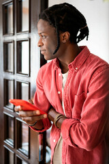 Thoughtful dreamy African American man with mobile phone in hand reflects on message received in internet chat. Young black guy stands indoors near wooden door and uses applications for online dating 