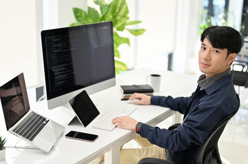 professional asian male programmer sits at his office desk, smiling at looking at the camera.