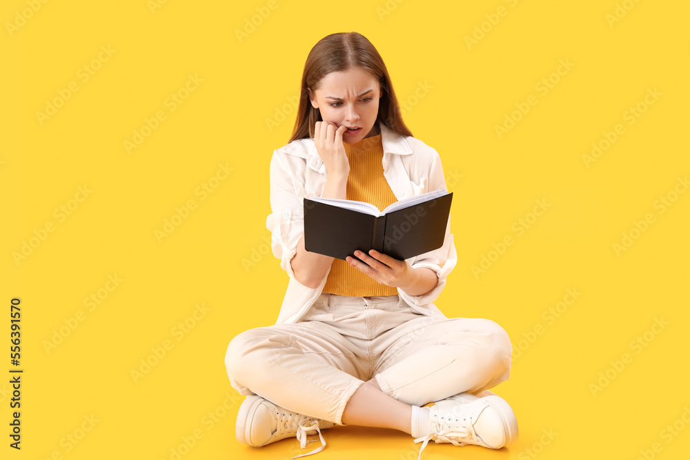 Young woman with book biting nails on yellow background