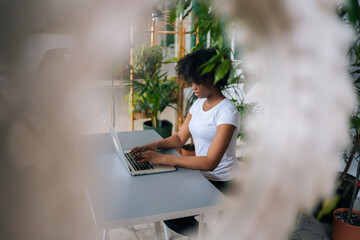 Side view of African-American female freelancer designer creating new fresh ideas, working on laptop computer in home office room with biophilic interior design, on background of green plants.