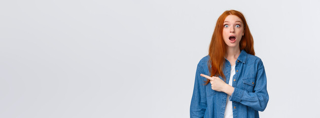 Wow so awesome, check this out. Impressed speechless and fascinated redhead female student drop jaw, looking astounded with popped eyes, pointing finger left, standing white background