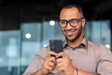 Close-up of smiling African-American businessman using phone typing messages and browsing internet pages, man in shirt and glasses smiling happily inside office holding smartphone.