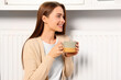 © New Africa - Woman holding cup with hot drink near heating radiator indoors