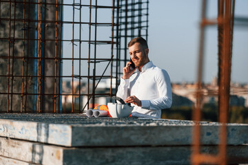 Wall Mural - Standing and talking by phone. Businessman in formal clothes is on the construction site at daytime