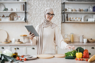 Wall Mural - Happy muslim woman in hijab cooking healthy food pointing on vegetables lying on counter while reading recipe on digital tablet in modern kitchen at home. Preparing vegetable salad.