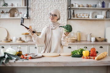 Wall Mural - Happy young muslim woman using smartphone in selfie stick standing in kitchen in the morning Happy female blogger holding broccoli speaking with followers in social media about preparing healthy salad