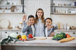 © sofiko14 - Young caucasian mother and two african sons waving hands preparing salad with fresh vegetables, hugging and smiling looking at the camera while cooking in kitchen. Healthy food, multinational family