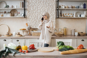 Portrait of young beautiful muslim woman in hijab standing at bright modern kitchen, using phone during call with her friends while cooking vegan salad from fresh vegetables at home.