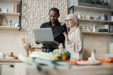 Wall Mural - Young wife and husband looking at online recipe on a digital laptop pc. Attractive muslim female and african male surfing the internet for recipes while making vegetarian salad in bright kitchen.