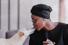 Close Up Shoot Of Young African Woman In Black Traditional Clothes Kissing Her  White Dog At Home. Adorable Moments Of Humans And Pets Friendship. Brazilian Female Plays With Puppy. Cute Animals.