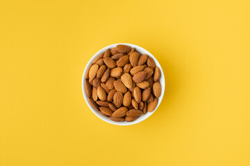 Wall Mural - Dried almonds in a white porcelain bowl on the yellow background. Nuts stacked together randomly with a copy space for a free text. Healthy nutrition concept. Flat lay