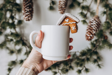 Blank white cup in hand with gingerbread house on Christmas decoration bokeh background. Mockup white cup.