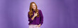 Glamour and stylish feminine woman with long natural red hair in purple sweater holding hand in pocket as pointing at upper left corner showing place she does hairstyle or makeup over violet wall