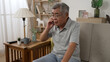 © PRPicturesProduction - senior man sitting on home sofa and answering call on cellphone during retirement time. boring elderly male hanging up after end the conversation on mobile phone while relax on couch in living room