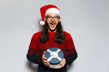 Wall Mural - Studio portrait of handsome laughing man with round gift box of blue color in hands. Holding round gift box. Wearing Santa Claus hat. Christmas concept. White background.