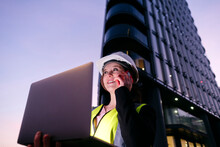Smiling Engineer Holding Laptop Talking On Mobile Phone In Front Of Building