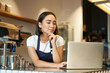 Leinwanddruck Bild - Portrait of smiling korean woman, barista in coffee shop, standing at counter with laptop, smiling and looking confident, self-employed female entrepreneur in her own coffee shop