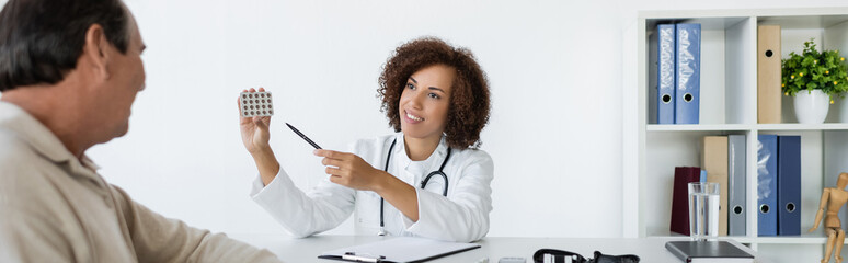 Positive african american doctor pointing at pills near mature man with diabetes in clinic, banner