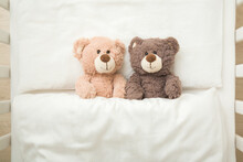 Two Adorable Smiling Light And Dark Brown Teddy Bears Lying Down On Pillow And Sheet Under Blanket In Baby Crib. Twins Concept. Top Down View. Closeup.