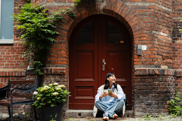 Wall Mural - Smiling asian girl using mobile phone and drinking coffee while sitting outdoors