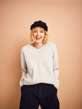 Studio Portrait Of A Young Woman Wearing A Grey Knit Sweater And Black Pants With Black Gatsby-style Hat Against A Brown Background; Studio