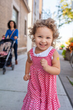 Preschooler Girl Running Down A Sidewalk In Her Red And White Gingham Dress And Looking Into The Camera With Her Mother And A Stroller In The Background; Toronto, Ontario, Canada