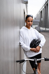 Wall Mural - Pretty african american woman in sunglasses holding helmet and looking at camera near bike in city 