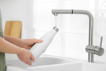 Sticker - Woman pouring fresh water from tap into thermo bottle in kitchen, closeup