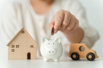 Close up of woman hand putting coin into White piggy bank for saving money, toy house and car on table, saving money and financial concept