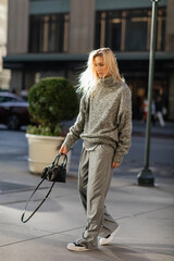 full length of stylish young woman with blonde hair holding handbag while walking in New York city in winter