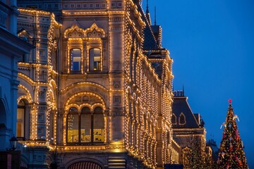 Wall Mural - a large illuminated building of GUM with lighted decorated giant christmas tree.Christmas decorations on a facade, big xmas tree with balls and presents, city night illumination.