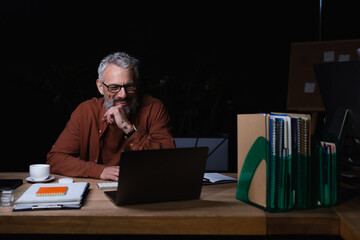 cheerful grey haired businessman looking at laptop while having video call at night in office