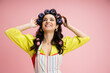 © LIGHTFIELD STUDIOS - cheerful young woman in yellow blouse and apron touching hair curlers and looking away isolated on pink