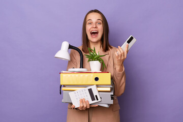 Image of extremely happy optimistic woman wearing beige jacket holding lot of documents folders isolated over purple background, holding mobile phone and expressing happiness, rejoicing, good news.