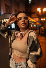 Fashionable Beautiful Stylish Hipster Girl With Pink Headphones In A Trendy Top With A Plaid Shirt And Jeans Shorts Wears Glasses And Walks In The Night City With Bokeh Lights