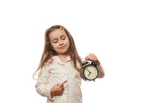 Falling Asleep, Sleepy Little Child Girl In White Pajamas, Posing With Closed Eyes, Pointing At Black Alarm Clock Showing The Bedtime, Isolated On White Background With Copy Advertising Space For Text