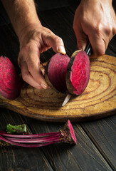 Sticker - Chef cuts with a knife red beets or beetroot on a wooden cutting board. Close-up of a cook hands with a knife on a kitchen black table. Peasant food