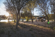 A Sunset Camping Scene In The Wild With A Camper Van In Between Trees On A Passing By River.