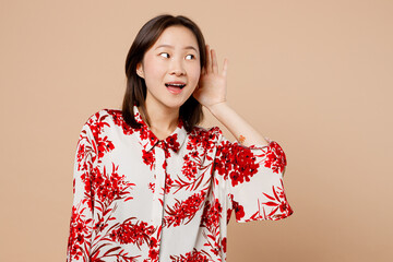 Wall Mural - Young curious nosy fun woman of Asian ethnicity wears red shirt try to hear you overhear listening intently isolated on plain pastel light beige background studio portrait. People lifestyle concept.