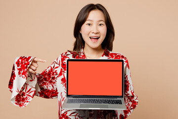 Wall Mural - Young smart IT woman of Asian ethnicity wears red shirt look camera hold use work point finger on laptop pc computer with blank screen workspace area isolated on plain pastel light beige background.