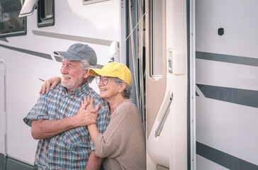 Lovely attractive senior couple on leisure trip standing outside a camper van looking away. Caucasian couple of retirees enjoying freedom and alternative life in motor home