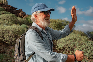 Sticker - Active smiling senior man outdoors enjoying healthy lifestyle goes trekking in mountain environment. Senior bearded male in hat and backpack walking with stick greeting someone