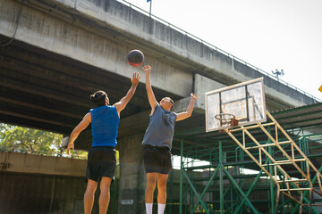 Wall Mural - Two man athlete playing streetball match shooting and defense basketball on outdoors court together in sunny day. Sportsman do sport training basketball at street court under highway in the city.