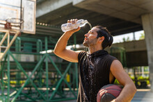Caucasian Sportsman Basketball Player Drinking And Pouring Water From A Bottle On His Face After Do Sport Training And Playing Streetball On Outdoors Court Under Highway In The City In Sunny Day.